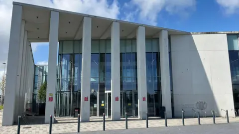 The picture shows the exterior of the court. It has a number of grey pillars resembling bars in front of a glass frontage. There is a solid grey wall with a royal crest.