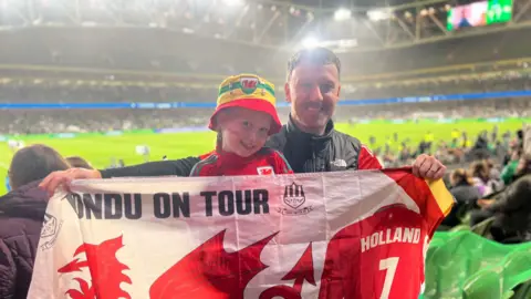 Joey Healey Joey Healey and Harlow supporting Wales successful  Dublin. They are holding a Wales flag, with the stadium down  them. Harlow has a Wales bucket chapeau  connected  and a Wales shot   apical  on.