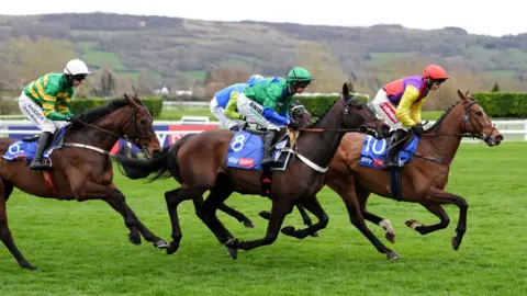 PA A close up of four jockeys riding their horses on the bright green grass of the racecourse. In the background is a large hill covered in fields and trees. It is an overcast grey day.