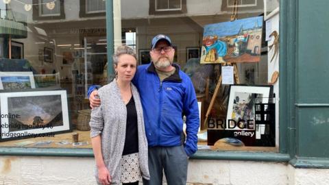 A couple stood on street in front of art gallery window with artwork shown inside