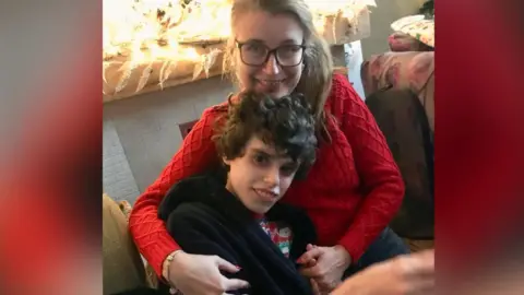 A woman with dark blonde hair and glasses wearing a red jumper hugs her son. He is wearing a Christmas top and a navy blue dressing gown. They are both smiling at the camera.