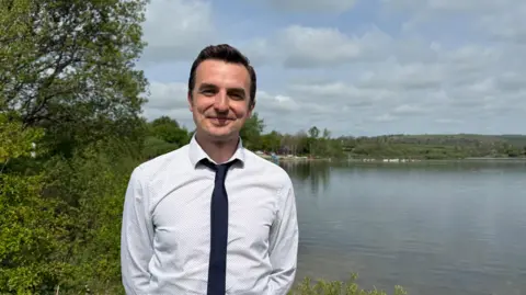 A man standing in front of Carsington Water in Derbyshire. He wears a white shirt with a black tie with his collar loosened. He has short dark hair and slightly smiles at the camera. Behind him is a big expanse of water, lined with green bushes.