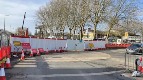 Road signs and barriers on the approach to the Botley Road from the side of the station. There is also a sign for diversion of traffic. There is one car on the road. The rail station can be seen in the distance.