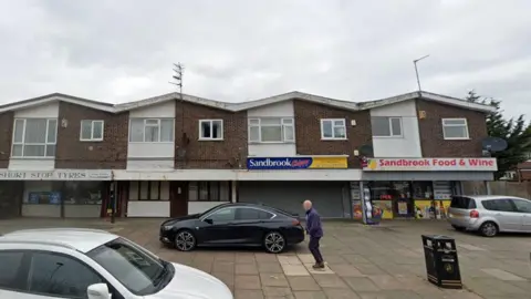 Google Homes and businesses on Sandbrook Road, Ainsdale. A number of cars are parked outside the properties and a man whose face is blurred is walking on the pavement.