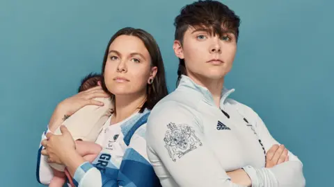 David Vintiner Two women stand side by side. The woman on the right has straight brown hair and she is holding a baby. The woman on the right has short brown hair. They are both wearing long-sleeved sports tops.