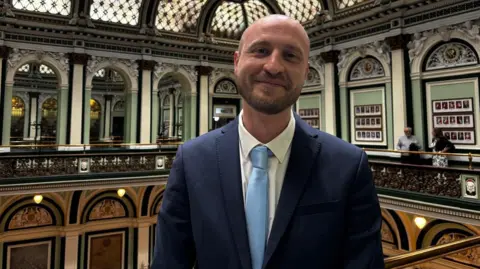 Gemma Dillon/BBC A man wearing a blue suit and blue tie smiling in a council chambers, he is bald with a beard 