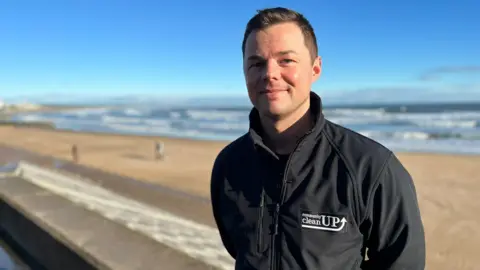 Man smiling at the camera wearing black jacket with community cleanUP logo on a beach in front of the sea.