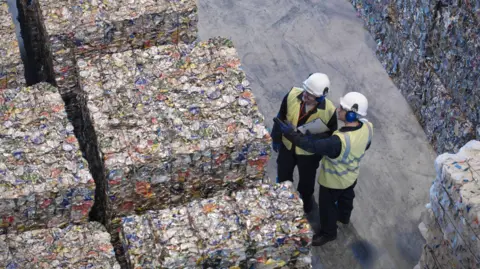 A recycling plant with blocks of waste lined up and two men in high visibility jackets, wearing hard hats, inspecting them.