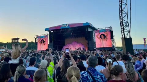 Festivalgoers watch Olivia Dean perform in the background. There is a floral design on the stage and the singer is wearing a pink dress. 