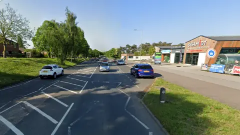 Google Cars travelling along Hall Road. A grass bank and trees line one side of the street, a retail park with wood-clad buildings lines the other