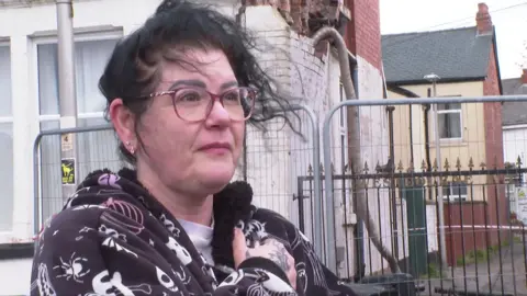 A woman in a brown patterned dressing gown and pyjamas and with tied back brown hair and glasses looks concerned as she is interviewed while standing in front of a house. A section of the side wall of the house is leaning and bricks are missing.