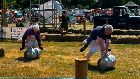 People hunch over grey metallic barrels and push them over grass. They are wearing sheep outfits around their legs.