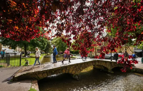 PA Bridge over the river Windrush in Bourton on the Water. Three people are crossing the bridge, with some red autumn leaves partly blocking the view of the river in the upper foreground. 