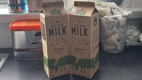 BBC Two cartons of 2.5% fat reduced milk on the counter in a kitchen with coffee and tea bags behind and a window. The cartons are light brown and have writing on them including "MILK 2.5%" on them. There is also images of green cows on the cartons. 