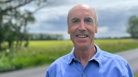 Head and shoulders image of Paul Boothman. Paul is wearing a blue shirt and is standing in front of a field and a road.