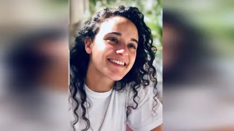 Mandy Damari Emily Damari, a young woman with long dark, curly hair, smiles while sitting in the sunshine, wearing a white T-shirt 