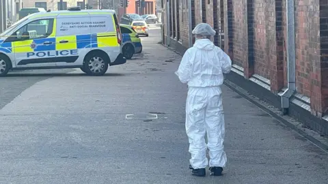 BBC A man wearing a white forensics suit is stood in a walkway with his back to the camera. Ahead of him is a police van and behind the van, two police cars can be seen. 