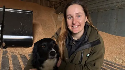 Anna Jackson is wearing a green jacket and kneeling next to her black and white boarder collie dog. They are in a grain store and a large pile of grain can be seen behind them.