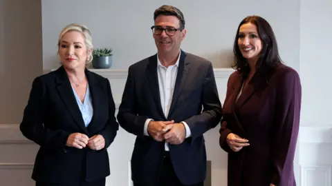 Reuters Britain's Prime Minister Andy Burnham meets with Northern Ireland's First Minister Michelle O'Neill and Deputy First Minister Emma Little-Pengelly during a visit to Northern Ireland. Michelle (left) has blonde hair tied back, wearing a navy blazer and light blue blouse. Andy (middle) has dark has dark hair, wearing glasses, a navy blazer and white shirt holding his hands together. Emma (right) has long dark hair, wearing a dark purple blazer.