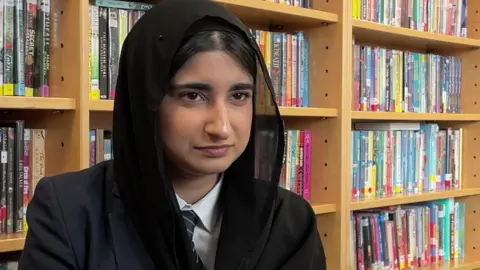 A teenage girl wearing a black headscarf sat next to a librarby shelf full of books