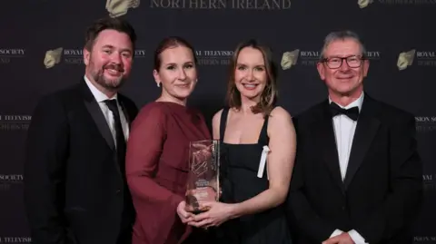 The BBC Newsline team - two men in black suits with white shirts and two female reporters. One is wearing a long sleeved maroon dress with her dark hair slicked back and the other is wearing a sleeveless black dress. They are holding the glass award.