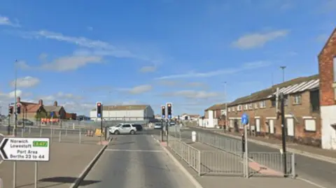 A pedestrian crossing. To the right, a pedestrian island separates the carriageway. To the left, a pedestrian island leads to another set of traffic lights further to the left. A white road sign pointing left states Norwich Lowestoft A47, Port Area Berths 23 to 44. Industrial buildings are in the background.