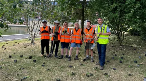 Andrew Spence Five people from the nursery are wearing orange high vis vests, and councillor Ross is wearing a yellow best and he holds a spade. Lots of 9ml pots of wildflowers are dotted around on the grass where they are to be planted.