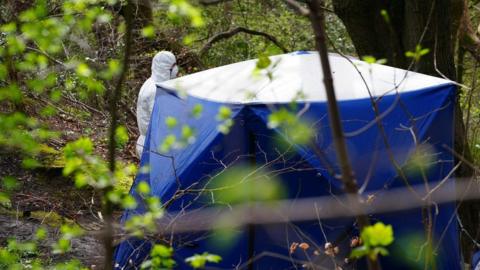 A crime scene investigator in a white forensic suit stands by a blue police tent
