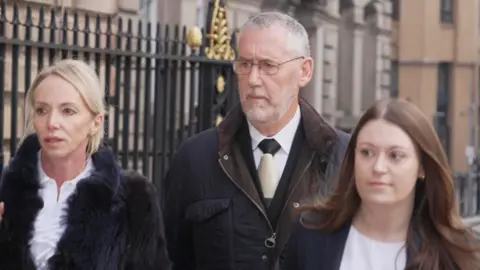 Ian Harvey, with short grey hair and a grey beard and wearing a dark blue coat, is pictured walking to Liverpool Town Hall flanked by two women, one blonde and wearing a fur style coat and one with long brown hair and wearing a dark blue blazer.