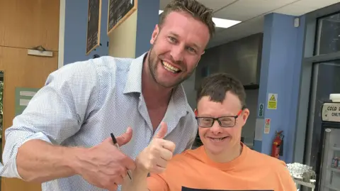 Steven Carruthers holding his thumb up, standing next to an adult with glasses wearing an orange T-shirt. Steven is wearing a blue patterned shirt, with stubble and short hair. They are in an office building. 