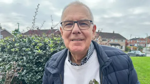 David Simcock has short grey hair and black glasses. He is wearing a white jumper with a blue and white shirt underneath, as well as a navy blue coat. He is wearing a prostate cancer UK pin, and a poppy.