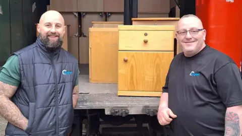 Two men, standing by an open lorry with lots of items inside, including cabinets and a red wheelie bin. The man on the left is wearing a blue gilet, with a green T-shirt, and the man on the right has on a black T-shirt. They are both smiling and looking straight at the camera. 