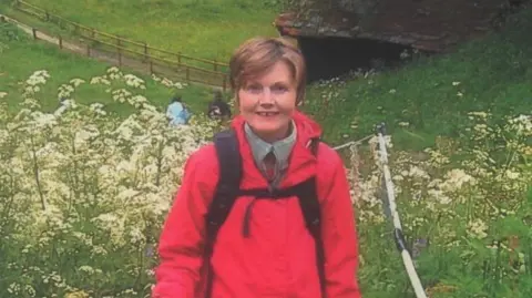 Alan Sheppard A woman with short brown hair is standing in a field. she is wearing a red jacket with a dark backpack.Behind her is a green field filled with white flowers, there is a stone building in the background and a path. 