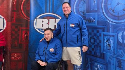 Two men, one in a wheelchair, both wear blue Bargain Hunt jumpers and pose in front of the Bargain Hunt signs smiling at the camera