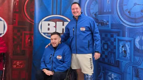 RAF Benevolent Fund Two men, one in a wheelchair, both wear blue Bargain Hunt jumpers and pose in front of the Bargain Hunt signs smiling at the camera