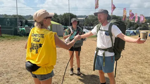A woman wearing a yellow t-shirt that reads "campsite crew" sprays water over a man's face. The man is wearing blue shorts, a white t shirt, hat and rucksack. He stands with his hands outstretched holding a coffee cup. A woman stands to his right in shorts and carrying bags. In the background is a stretched of browned grass and pink flags.