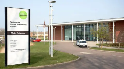 Amy Holmes/BBC A modern council building sits beside a curved driveway, with a tall sign in front reading: “Welcome to Central Bedfordshire Council” and “Main Entrance”. The area has grass, trees and a few parked vehicles under a clear sky.