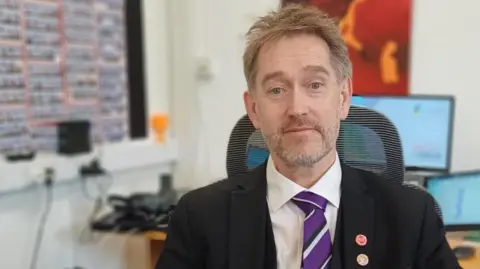 Debbie Tubby/BBC A man with light brown short hair and a greying beard. He is wearing a dark jacket over a white shift and striped tie. He is sitting down and behind him are computer screens. 