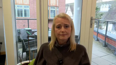 Justine has long long blond hair and wears a dark top. She stands in front of a window with terrace furniture in the background