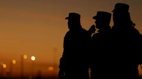 Silhouette of three Mexican soldiers at the US border. The sky in the background looks like it is sunset 