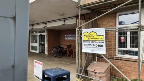Callum Brunsdon A hospital entrance. There is scaffolding to the side of the frame with the contractors sign on it.