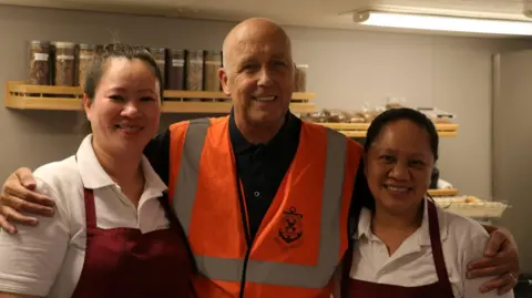 Paul Atkinson is standing with his arms around two women who are wearing aprons and smiling. He is wearing a high-vis jacket with the words Stella Maris on it 