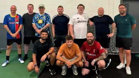 Forget-Me-Knot FC Ten men pose for a team photo in front of a football goal inside a sports hall. Seven men are standing while three others are crouched in front of them.