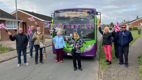 Bus passengers gather around a bus, waving Union flags