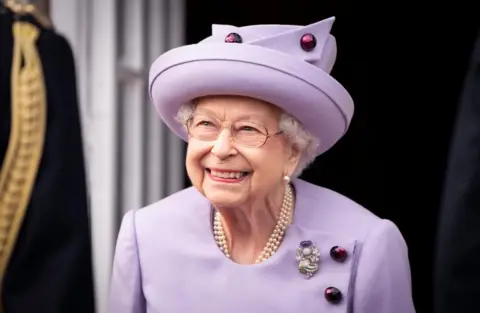 PA Queen Elizabeth II seen looking upwards out of the shot. She is wearing a pink dress and matching wide-brimmed hat. 