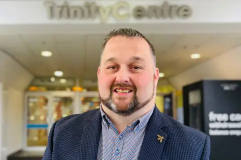 Kenny Bruce smiling at the camera, he is wearing a blue suit jacket and a patterned shirt, the words Trinity Centre are behind him on a sign.