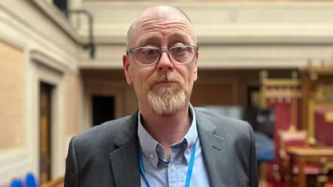 David McCaughey wears a grey jacket, blue shirt and glasses. He has grey hair and a beard.