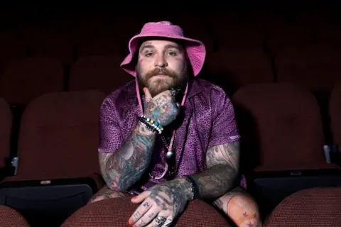 Chapman Byler Teddy Swims rests his head in his hand, while sitting in movie theater-style seating, wearing a pink bucket hat and a purple leopard-print shirt.