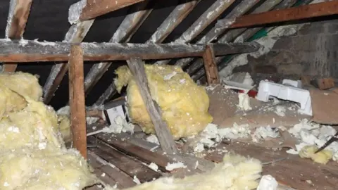 An attic space with wooden beams. There are two huge balls of insulation foam and the whole space is scattered with pieces of broken polystyrene. 