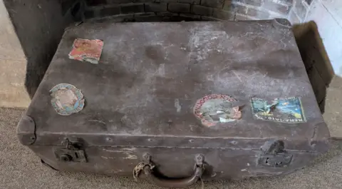A battered and dusty brown suitcase with four tattered stickers on it